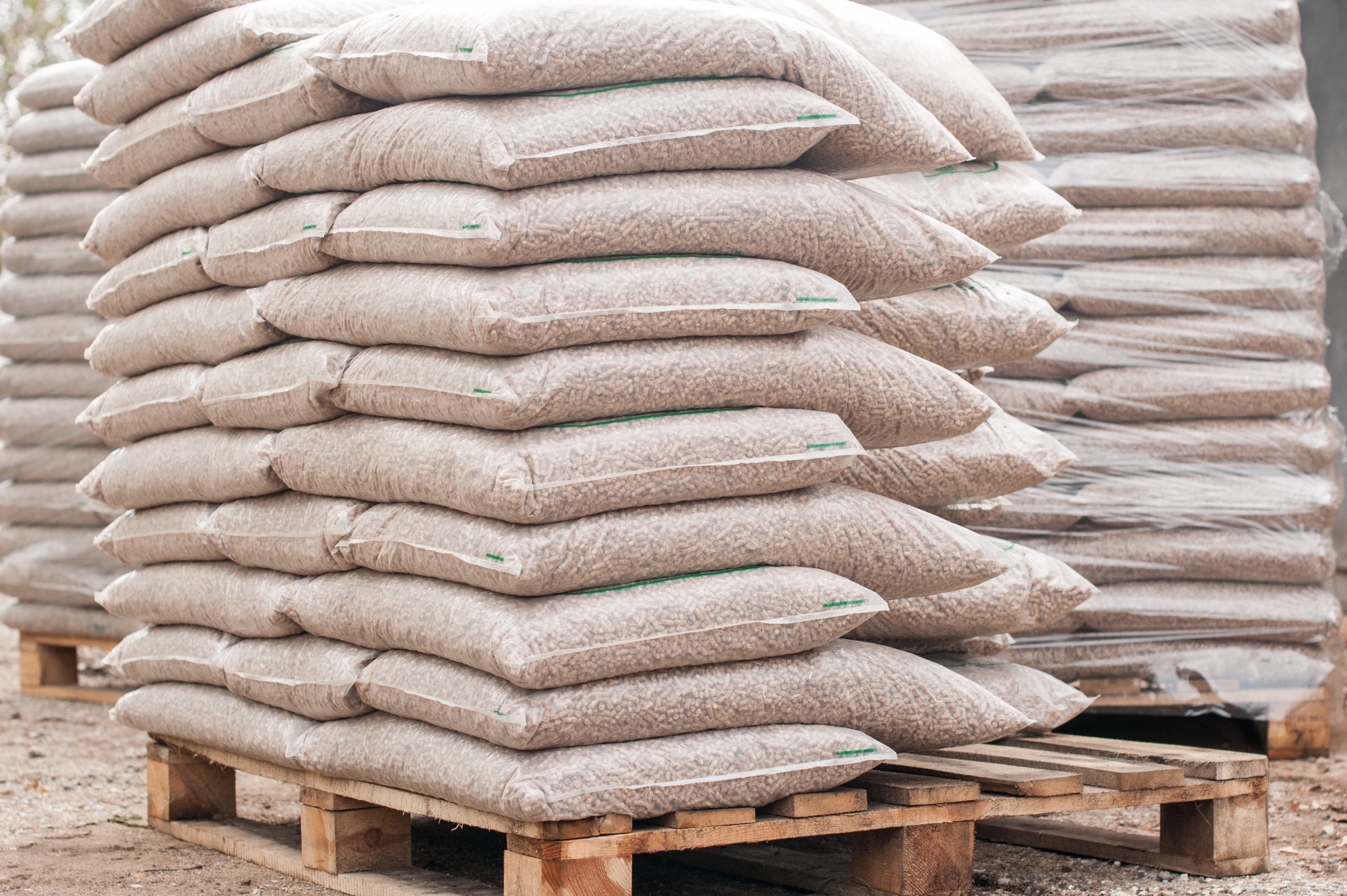 Ułożone piramidalnie worki z pelletem na drewnianych paletach, widoczne przezroczyste opakowanie i struktura pelletu, drugi stos worków owinięty folią stretch.