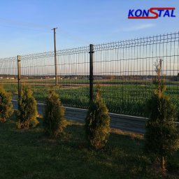Ogrodzenia Konstal - Nowoczesne, czarne ogrodzenie panelowe z podmurówką i rzędem młodych tuj, widok na pola i zabudowania w tle, logo firmy w górnym rogu.