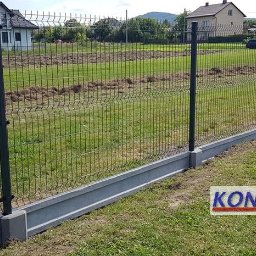 Ogrodzenia Konstal - Nowoczesne ogrodzenie panelowe z podmurówką betonową na tle zielonego trawnika i zabudowy jednorodzinnej, z widocznym logo producenta umieszczonym w prawym dolnym rogu.