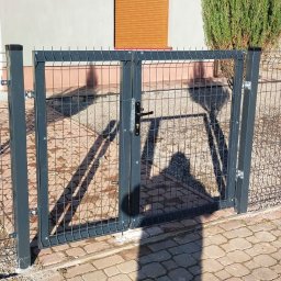 Ogrodzenia Konstal - Dwuskrzydłowa furtka z metalowej siatki z czarną klamką, wkomponowana w ogrodzenie panelowe, widoczna z zewnątrz posesji, z młodym drzewkiem iglastym po lewej stronie i ścieżką z kostki brukowej.