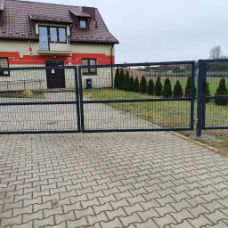 Ogrodzenia Konstal - Otwarta dwuskrzydłowa brama z paneli siatkowych w kolorze grafitowym, prowadząca na posesję z budynkiem administracyjnym i rzędem małych tuj.