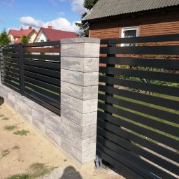 Ogrodzenia Konstal - Nowoczesna, pozioma, czarna brama przesuwna z wypełnieniem z płaskich profili, osadzona na murku z szarej kostki łupanej z widoczną fakturą, w tle domy jednorodzinne.