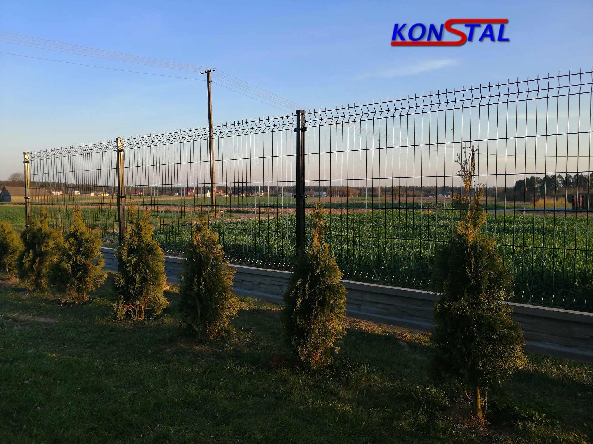 Nowoczesne, czarne ogrodzenie panelowe z podmurówką i rzędem młodych tuj, widok na pola i zabudowania w tle, logo firmy w górnym rogu.