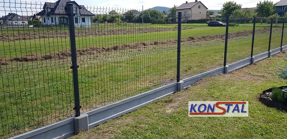 Nowoczesne ogrodzenie panelowe z podmurówką betonową na tle zielonego trawnika i zabudowy jednorodzinnej, z widocznym logo producenta umieszczonym w prawym dolnym rogu.