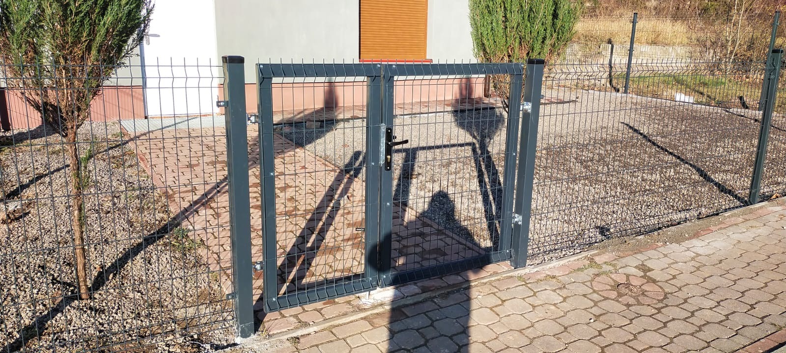 Dwuskrzydłowa furtka z metalowej siatki z czarną klamką, wkomponowana w ogrodzenie panelowe, widoczna z zewnątrz posesji, z młodym drzewkiem iglastym po lewej stronie i ścieżką z kostki brukowej.