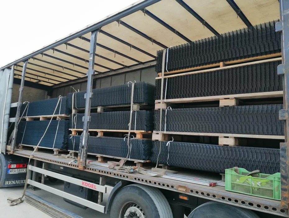 Ciężarówka załadowana paletami z czarnymi panelami ogrodzeniowymi, widoczne tablice rejestracyjne pojazdu i logo producenta naczepy.