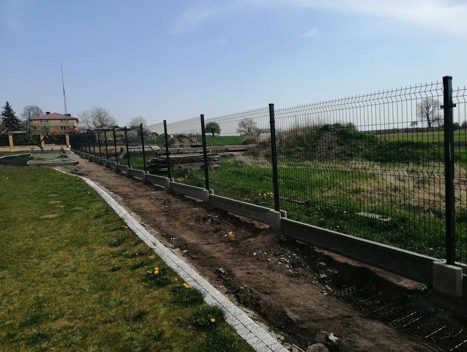 Długi odcinek nowego ogrodzenia z paneli z podmurówką betonową w trakcie montażu, widoczny wykop z siatką zbrojeniową, na trawniku żółte kwiaty.
