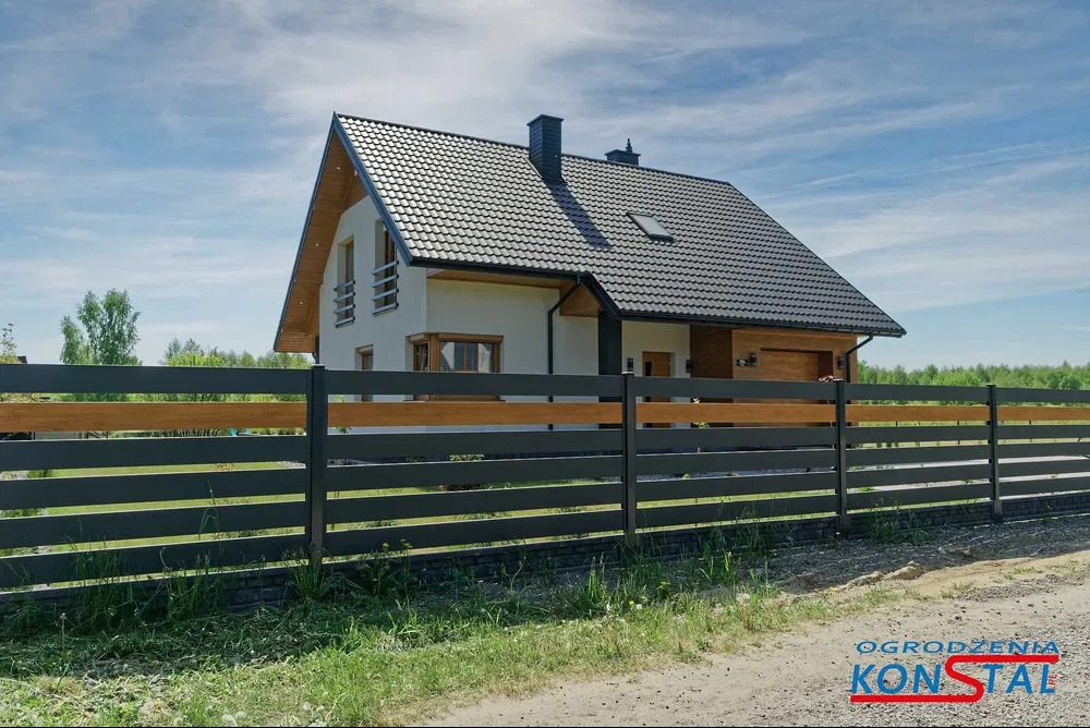 Nowoczesne ogrodzenie poziome z szarych, metalowych paneli z drewnianymi akcentami na tle domu jednorodzinnego w słoneczny dzień.