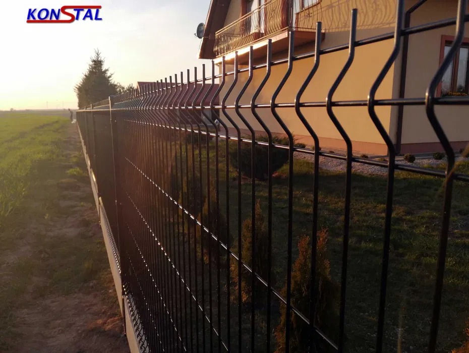 Czarne ogrodzenie panelowe z widocznym logo producenta KONSTAL na tle domu i zachodzącego słońca.