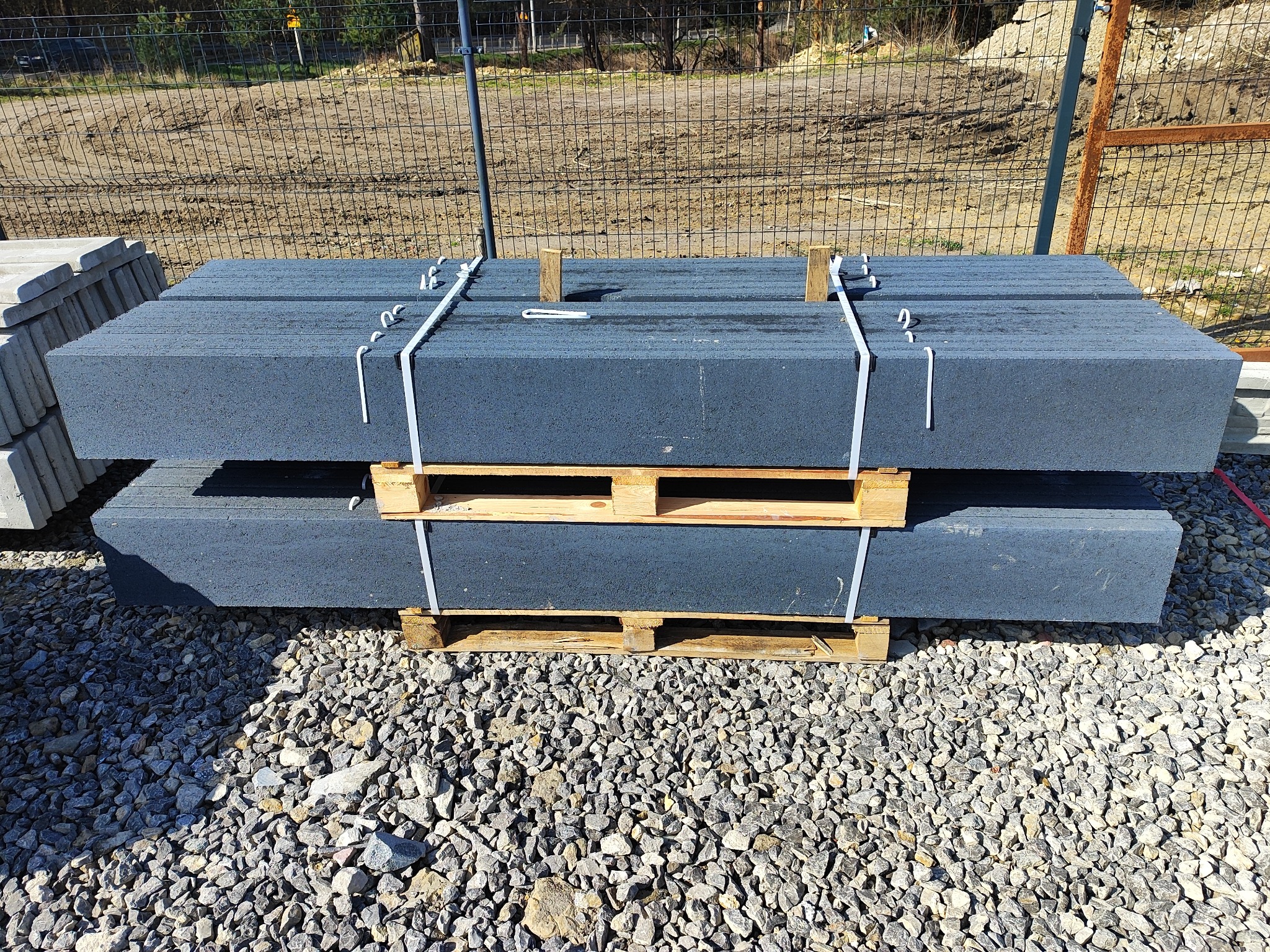 Paleta z betonowymi elementami ogrodzeniowymi w kolorze grafitowym, zabezpieczona taśmą, na tle żwirowego podłoża i fragmentu ogrodzenia panelowego.