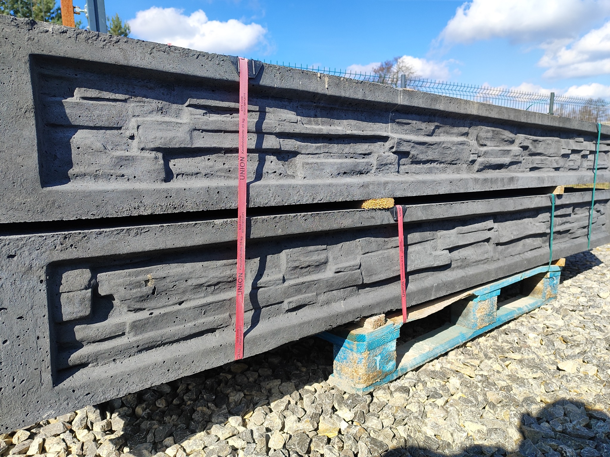 Szare, betonowe panele imitujące kamień, ułożone na niebieskiej palecie, zabezpieczone czerwonymi taśmami transportowymi, na tle żwirowego podłoża i fragmentu siatki ogrodzeniowej.