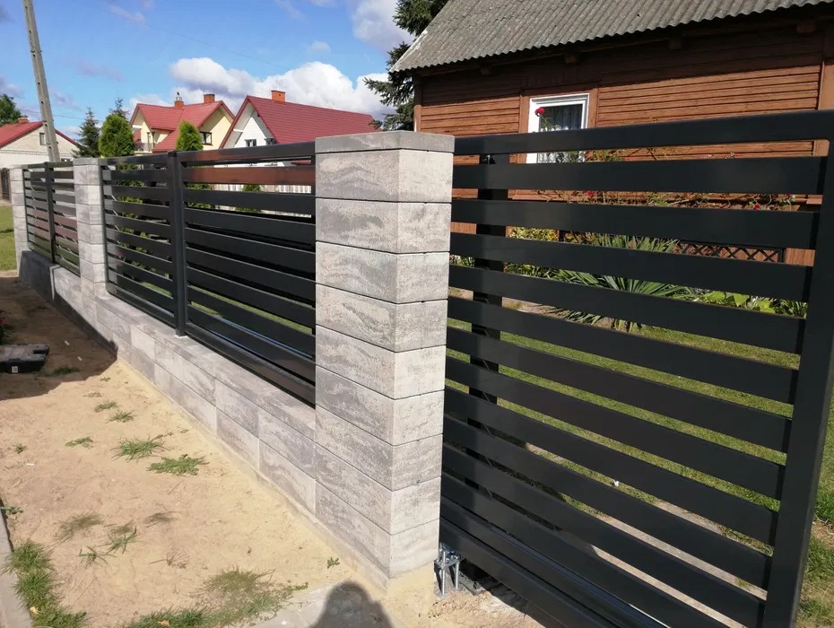 Nowoczesna, pozioma, czarna brama przesuwna z wypełnieniem z płaskich profili, osadzona na murku z szarej kostki łupanej z widoczną fakturą, w tle domy jednorodzinne.