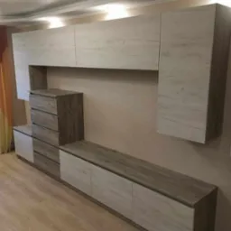 Zestaw mebli do salonu z szufladami i szafkami wiszącymi o teksturze jasnego drewna z widocznymi słojami, ustawiony pod sufitem podwieszanym z oświetleniem punktowym.