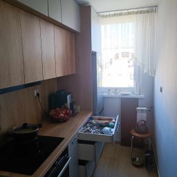 Gl meble meble na wymiar - Wnętrze kuchni z drewnianymi frontami szafek, czarną płytą indukcyjną i otwartą szufladą z przyborami kuchennymi, widok na okno z firanką i grzejnik.