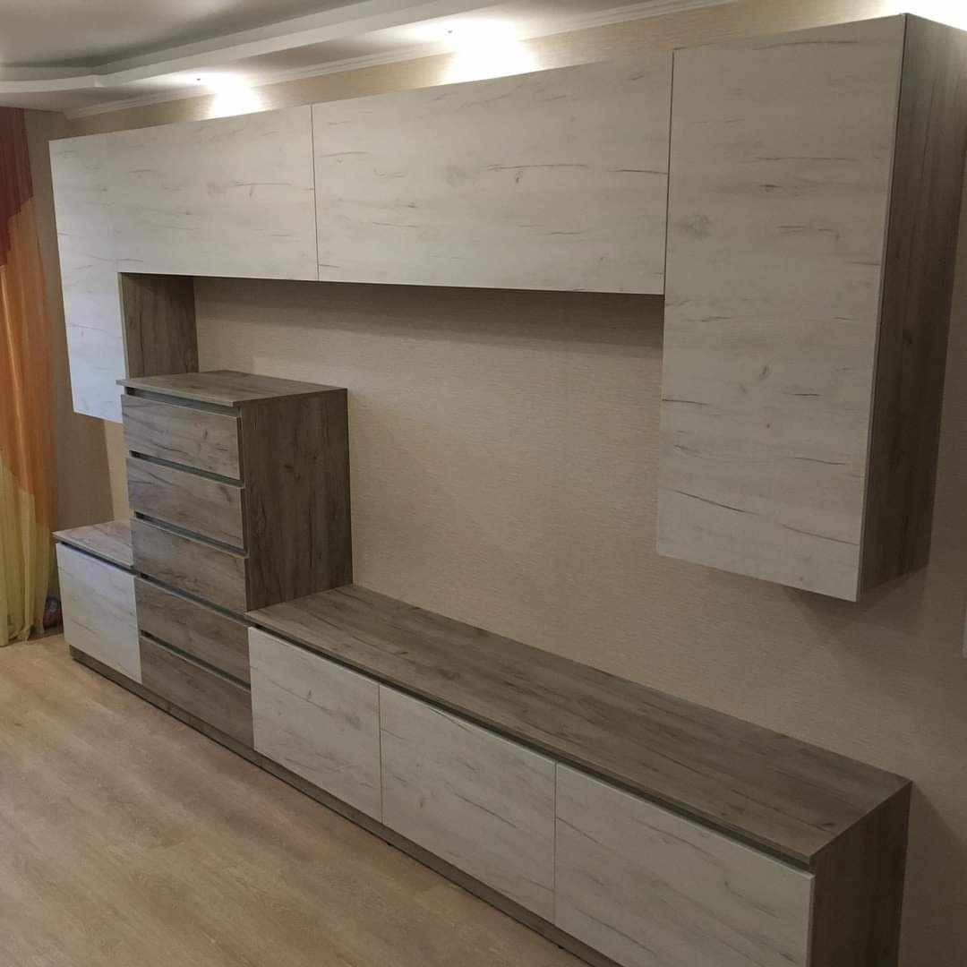 Zestaw mebli do salonu z szufladami i szafkami wiszącymi o teksturze jasnego drewna z widocznymi słojami, ustawiony pod sufitem podwieszanym z oświetleniem punktowym.