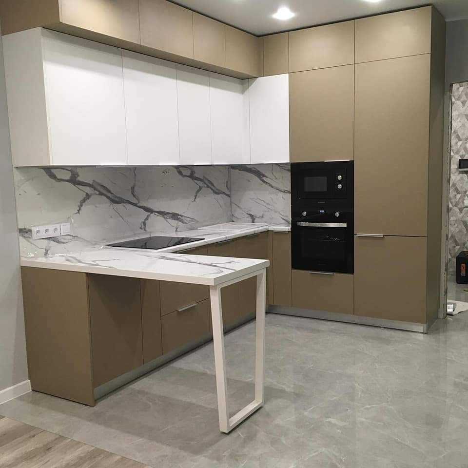Nowoczesna kuchnia z białymi szafkami górnymi i beżowymi dolnymi, blatem imitującym marmur i wyspą z białym stelażem, oświetlona światłem punktowym.