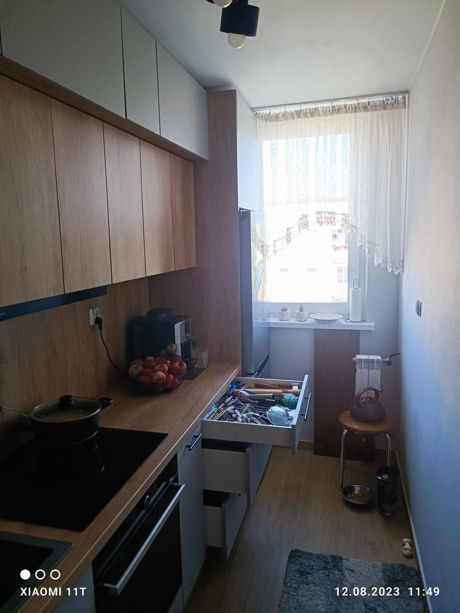 Wnętrze kuchni z drewnianymi frontami szafek, czarną płytą indukcyjną i otwartą szufladą z przyborami kuchennymi, widok na okno z firanką i grzejnik.