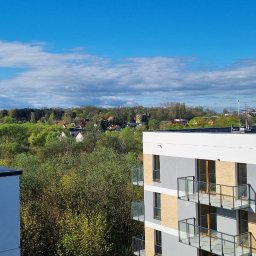 ADNIER S.C. - Widok z balkonu nowoczesnego apartamentowca w Krakowie na zieloną okolicę i błękitne niebo z chmurami.