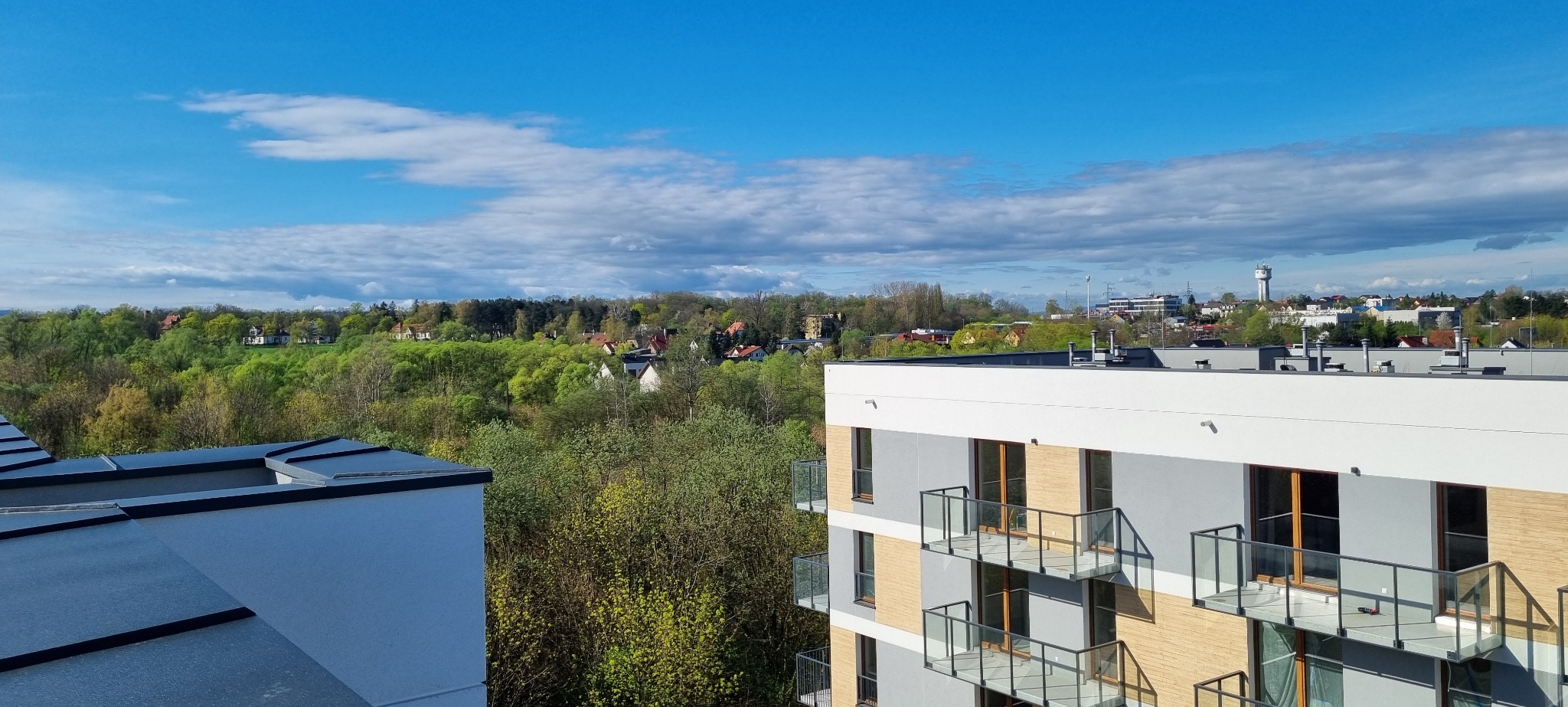 Widok z balkonu nowoczesnego apartamentowca w Krakowie na zieloną okolicę i błękitne niebo z chmurami.