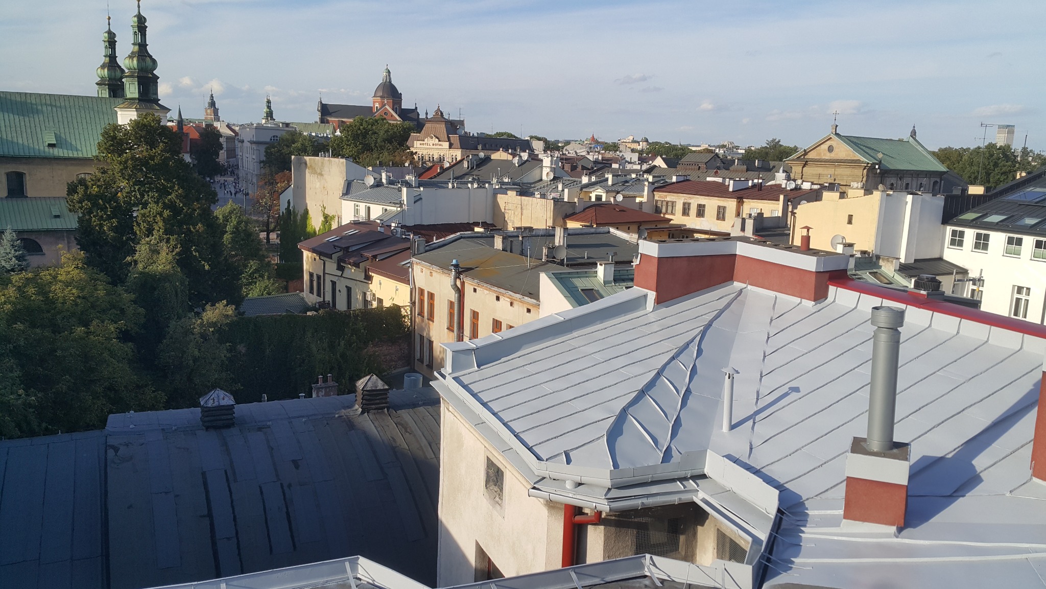 Panorama miasta z widokiem na dachy budynków, w tym nowo położony, jasnoszary dach z elementami w kolorze ceglastym, na tle zabytkowej architektury i zieleni drzew.