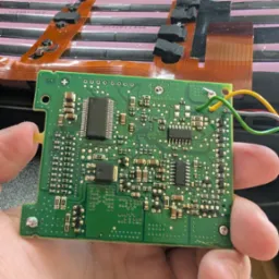 Zielona płytka drukowana z układami scalonymi, trzymana w dłoni, z widocznymi przewodami. W tle narzędzia i elementy elektroniczne.