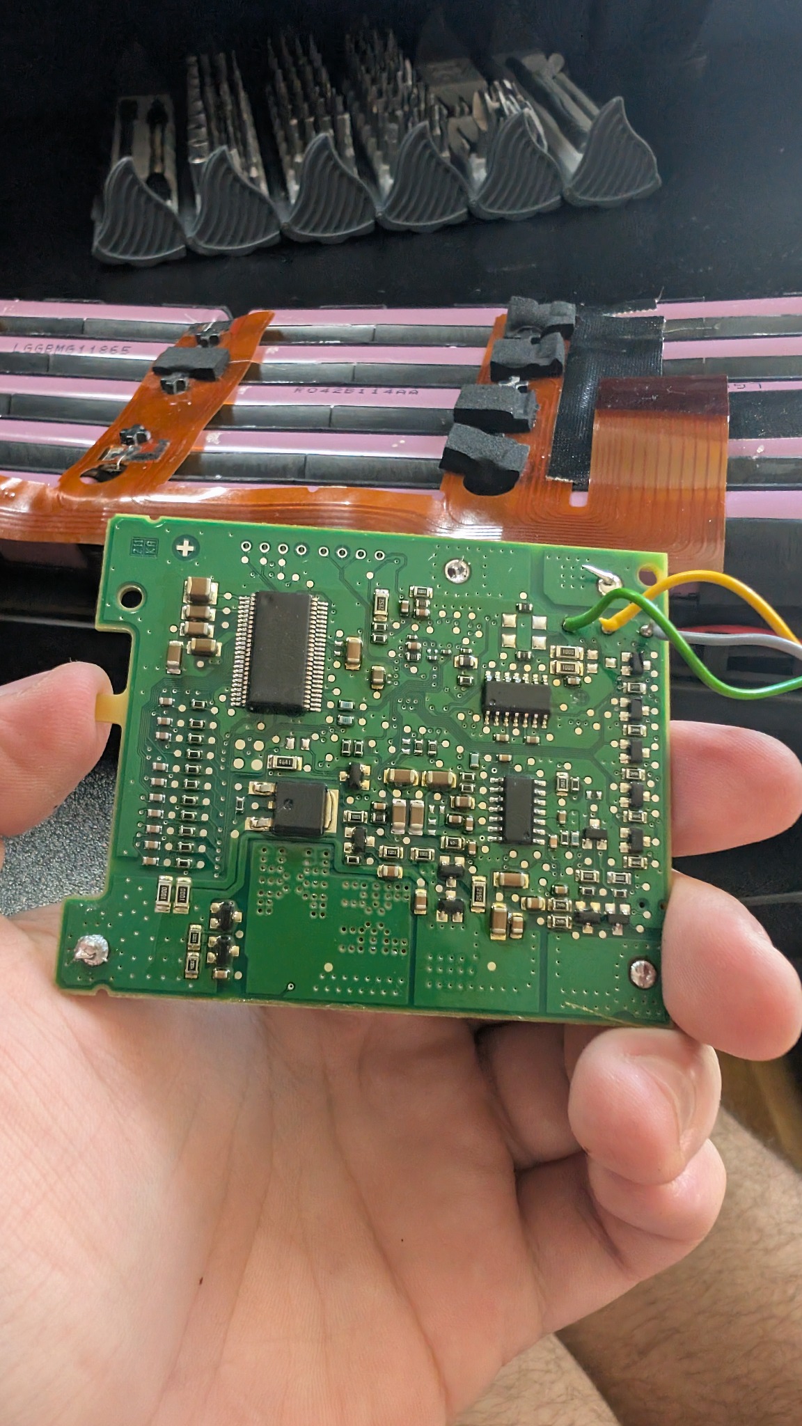 Zielona płytka drukowana z układami scalonymi, trzymana w dłoni, z widocznymi przewodami. W tle narzędzia i elementy elektroniczne.