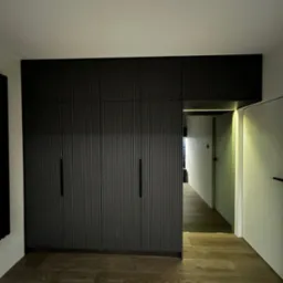 Nowoczesna, ciemna szafa wnękowa z ryflowanymi frontami, zintegrowana z oświetleniem nad przejściem do korytarza, na tle drewnianej podłogi.