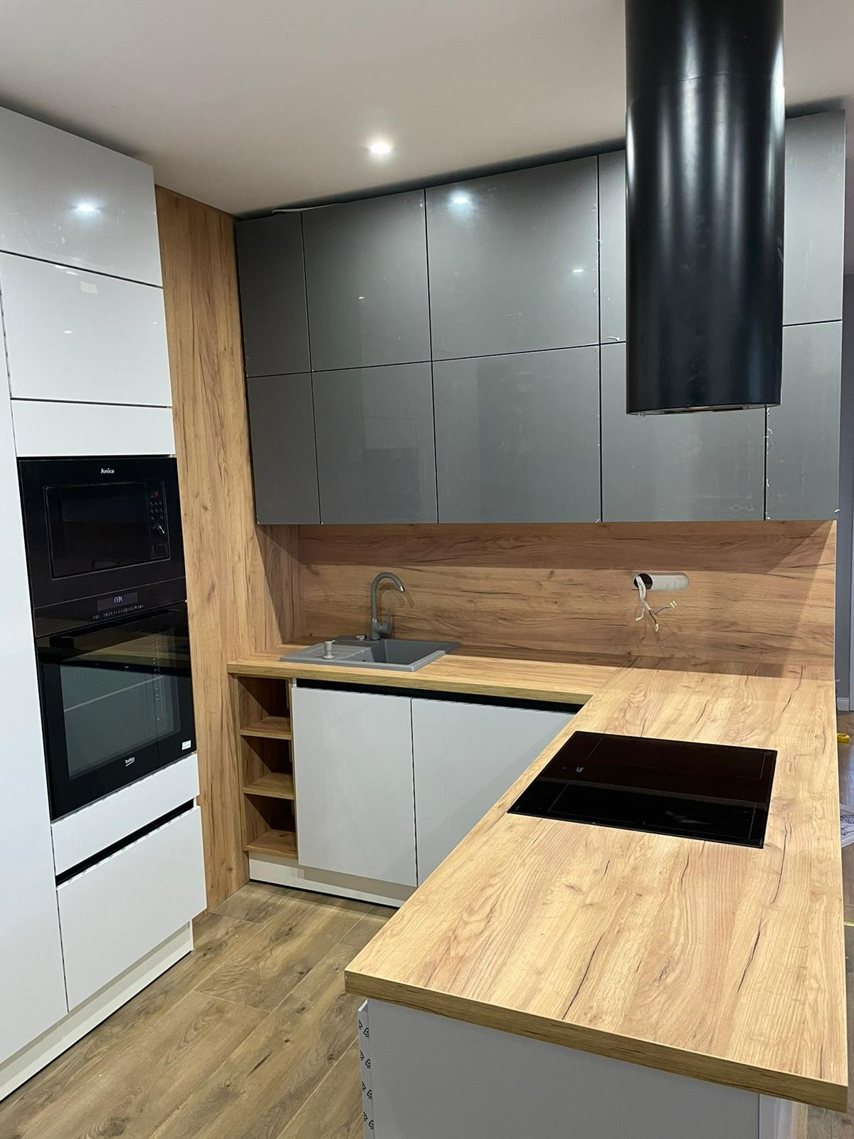 Nowoczesna kuchnia z szarymi szafkami wiszącymi, drewnianym blatem i okładziną ścienną, czarnym okapem oraz białymi szafkami dolnymi i piekarnikiem Amica.