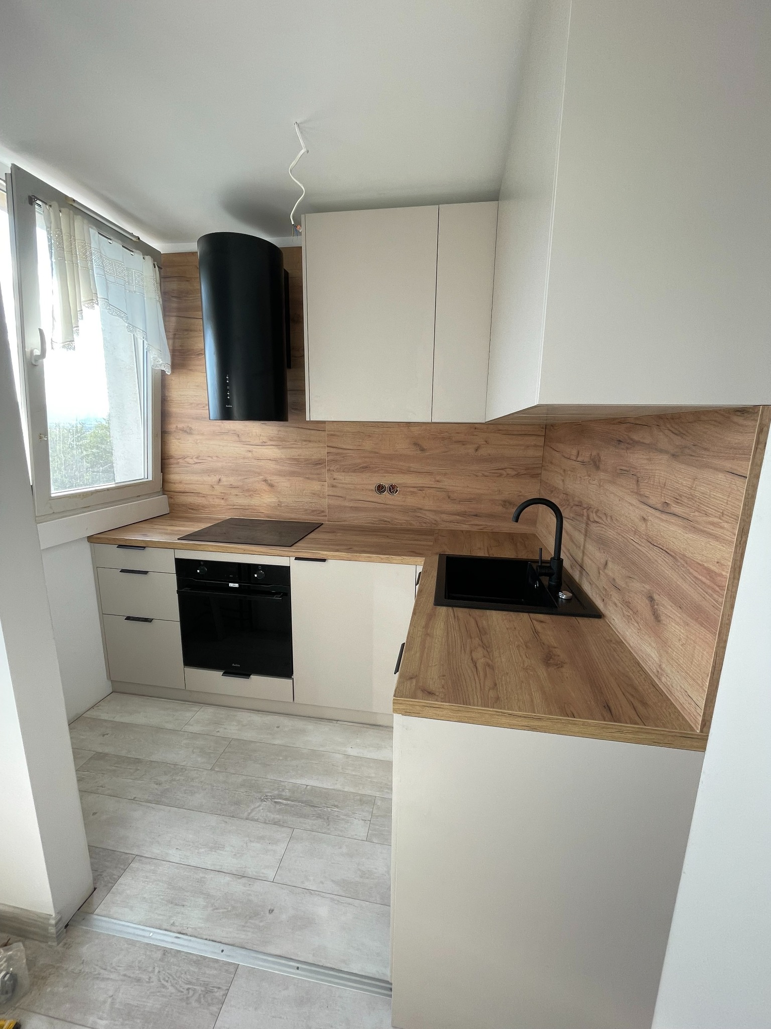 Niewielka kuchnia w bloku z jasnymi szafkami i drewnopodobnym blatem, czarny okap cylindryczny i czarny zlew, widoczne niedokończone prace wykończeniowe.