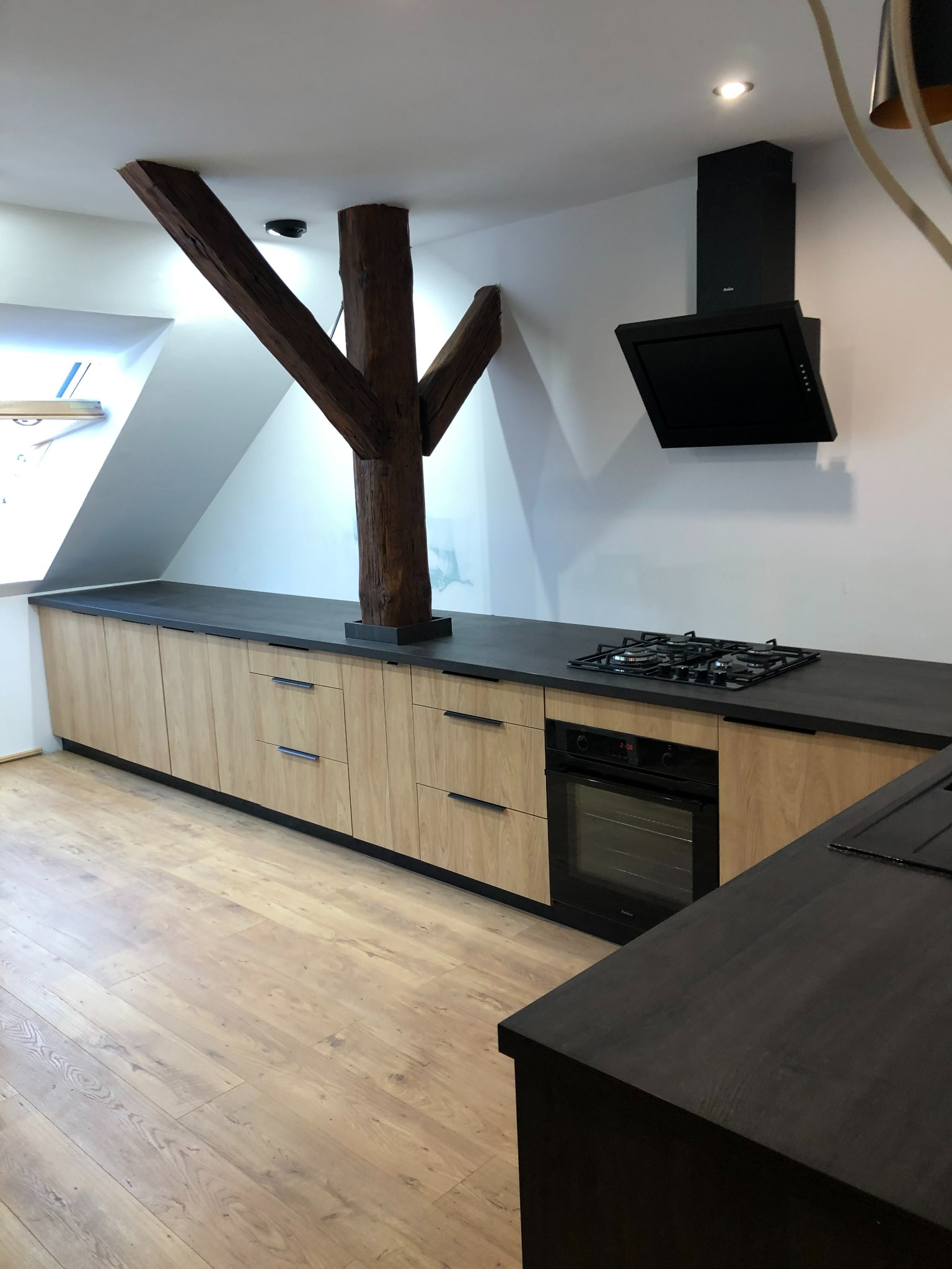 Nowoczesna kuchnia z drewnianymi frontami szafek i ciemnym blatem, integrująca istniejącą, drewnianą kolumnę konstrukcyjną w zabudowie.