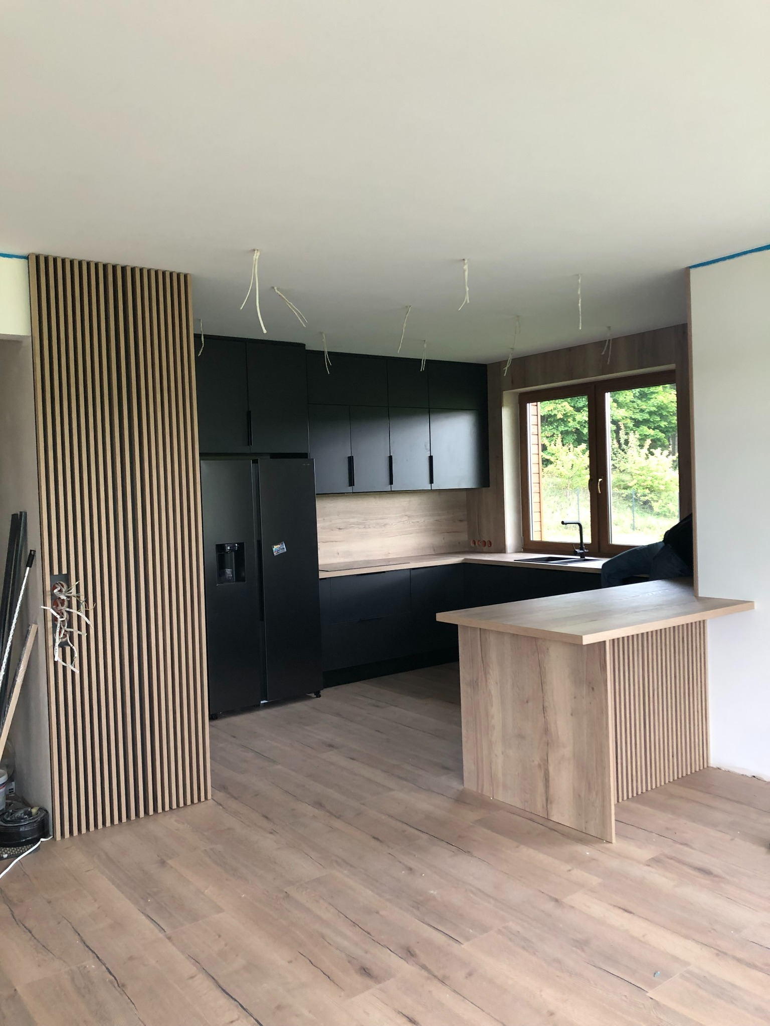 Nowoczesna kuchnia w trakcie montażu: czarne szafki, drewniane blaty, lamelowa ściana, widoczne przewody elektryczne.