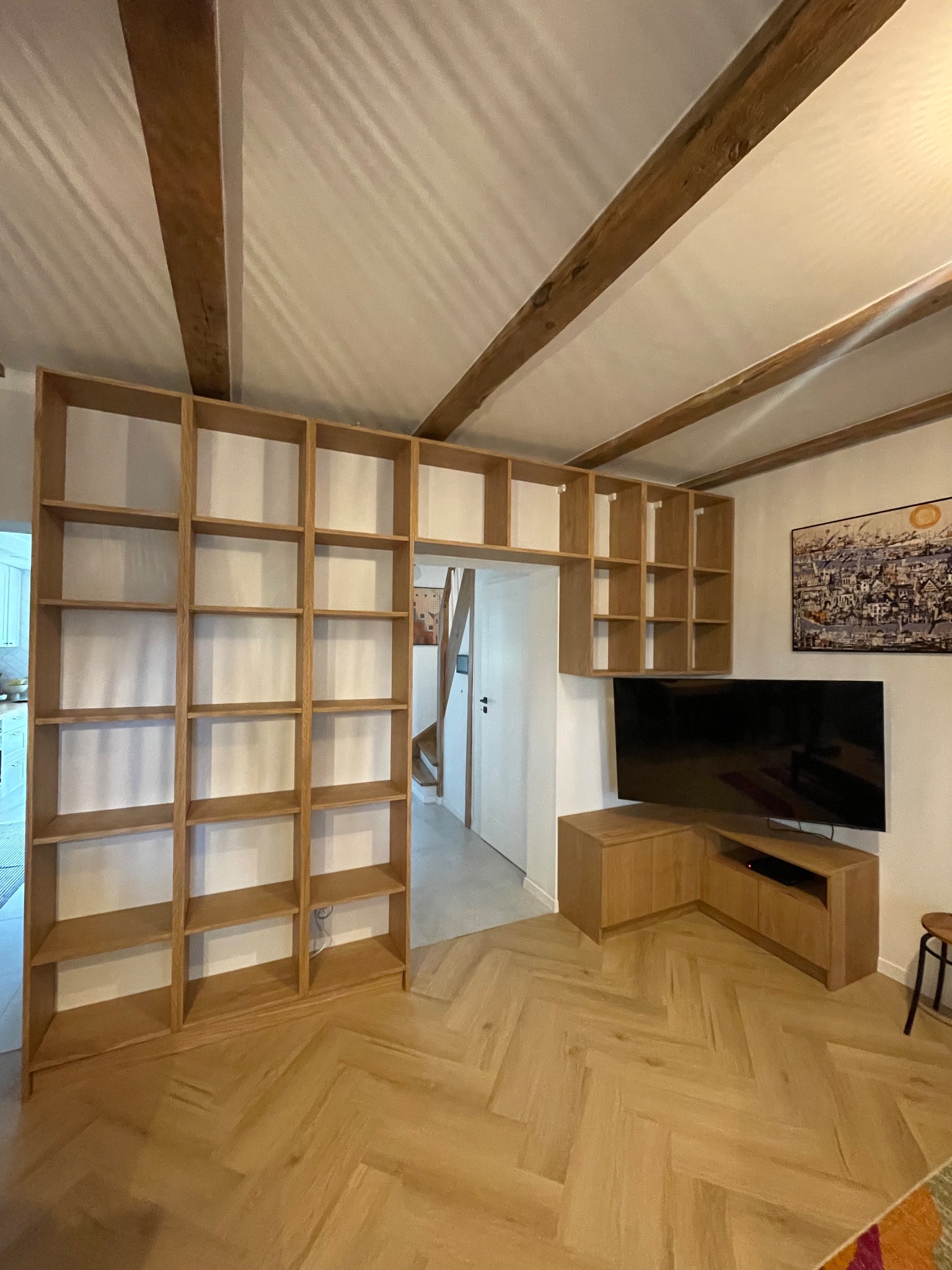 Drewniana zabudowa salonu z regałem na książki, szafką RTV i telewizorem, podwieszona szafka nad przejściem, parkiet w jodełkę, drewniane belki stropowe.