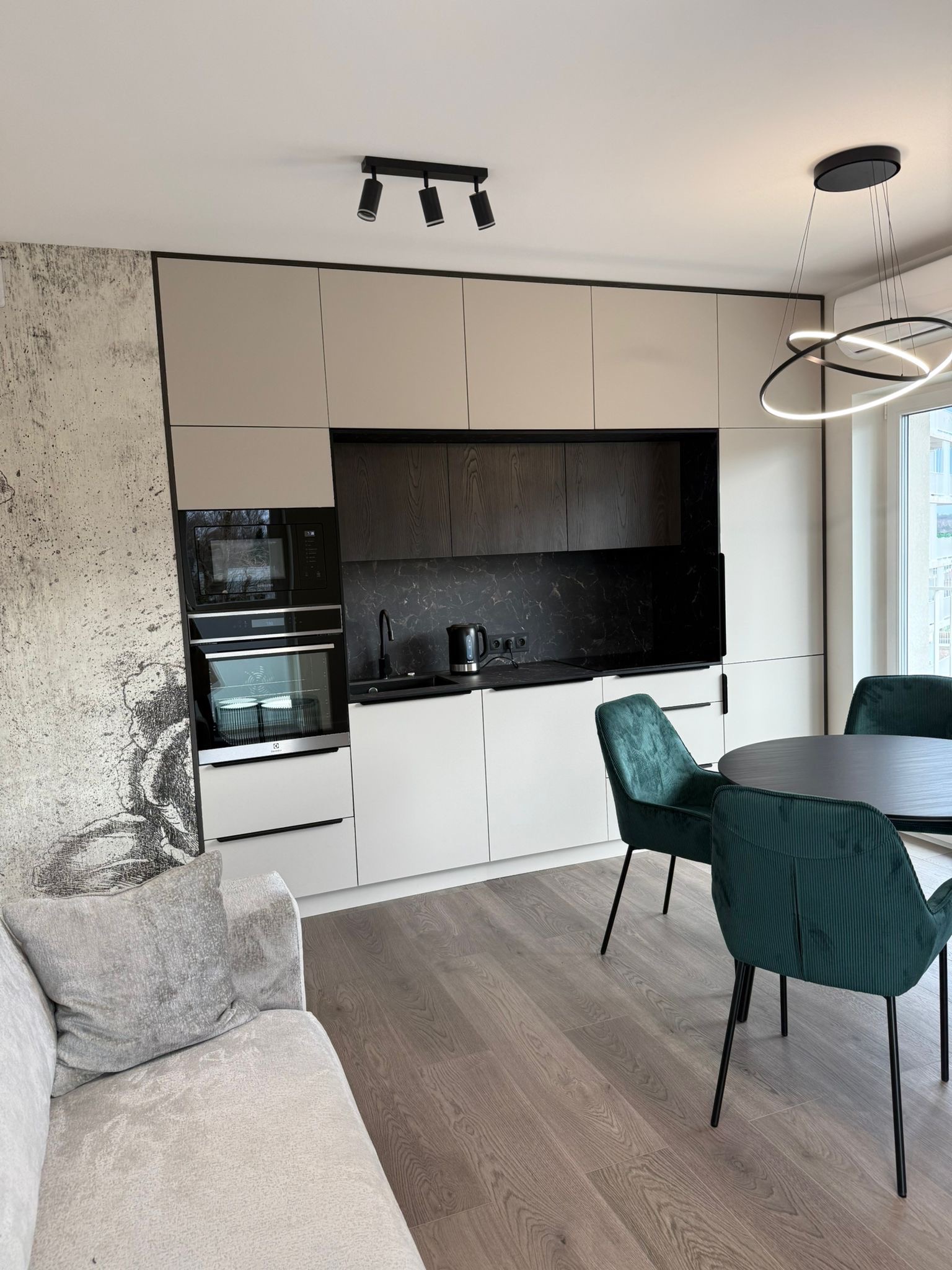 Nowoczesna kuchnia z zabudową w odcieniach beżu i czerni, widoczny fragment salonu z szarą sofą i okrągłym stołem z zielonymi krzesłami, oświetlenie sufitowe punktowe i designerska lampa wisząca.