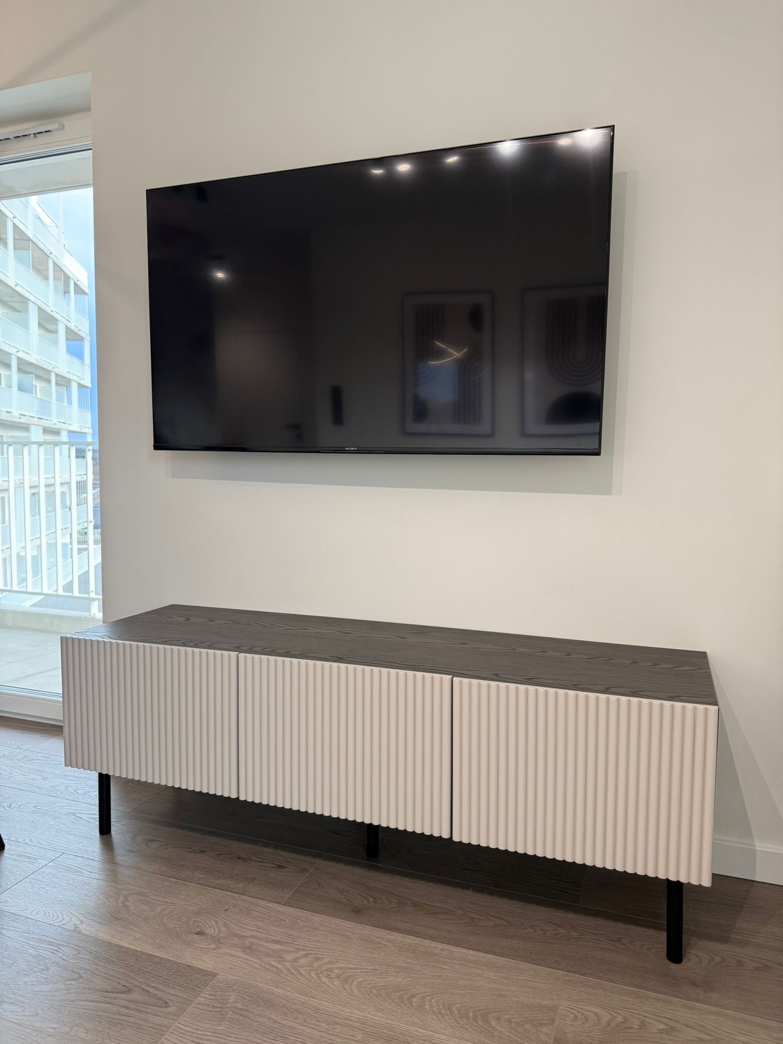 Nowoczesna szafka RTV z ryflowanymi frontami w kolorze złamanej bieli i blatem z ciemnego drewna, ustawiona pod wiszącym telewizorem w minimalistycznym salonie z widokiem na balkon.