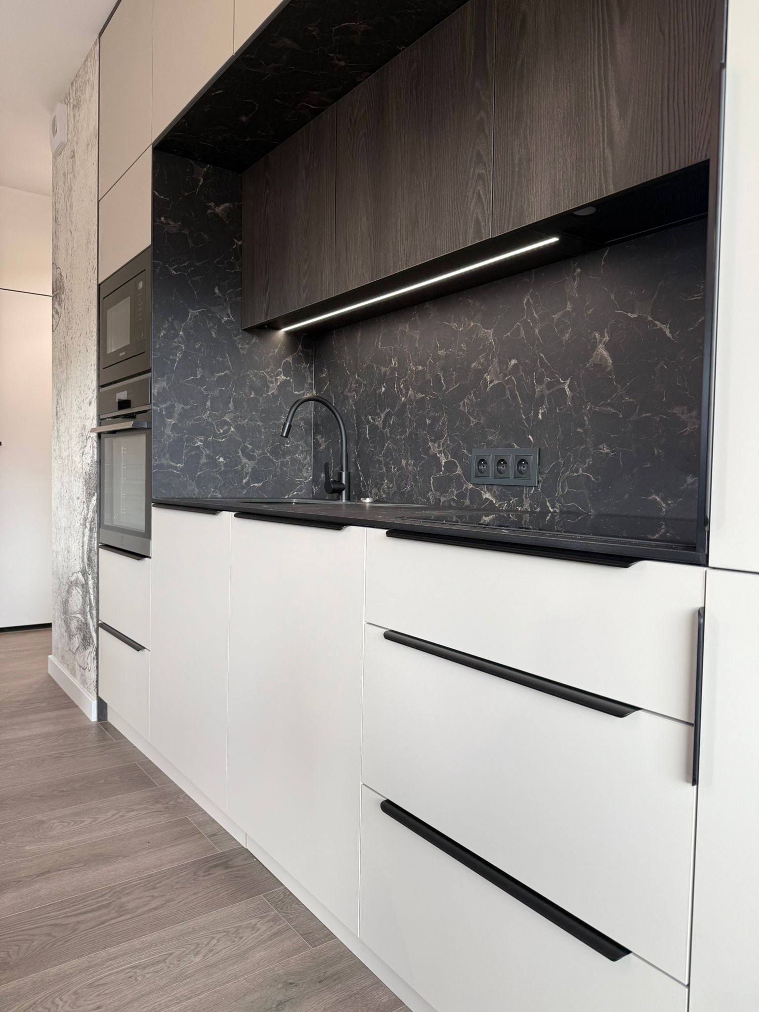Nowoczesna kuchnia z białymi frontami szafek, czarnym blatem imitującym marmur, ciemnobrązowymi szafkami górnymi i podświetleniem LED pod szafkami.
