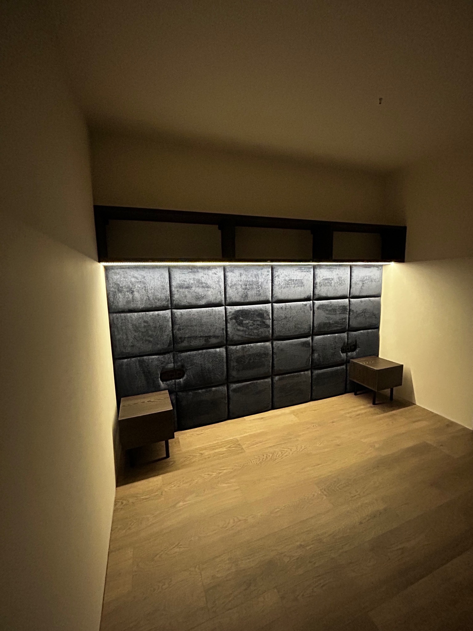 Tapicerowany panel ścienny w sypialni z oświetleniem LED, dwie drewniane szafki nocne na cienkich nóżkach, parkiet na podłodze, ciemne półki nad panelem.