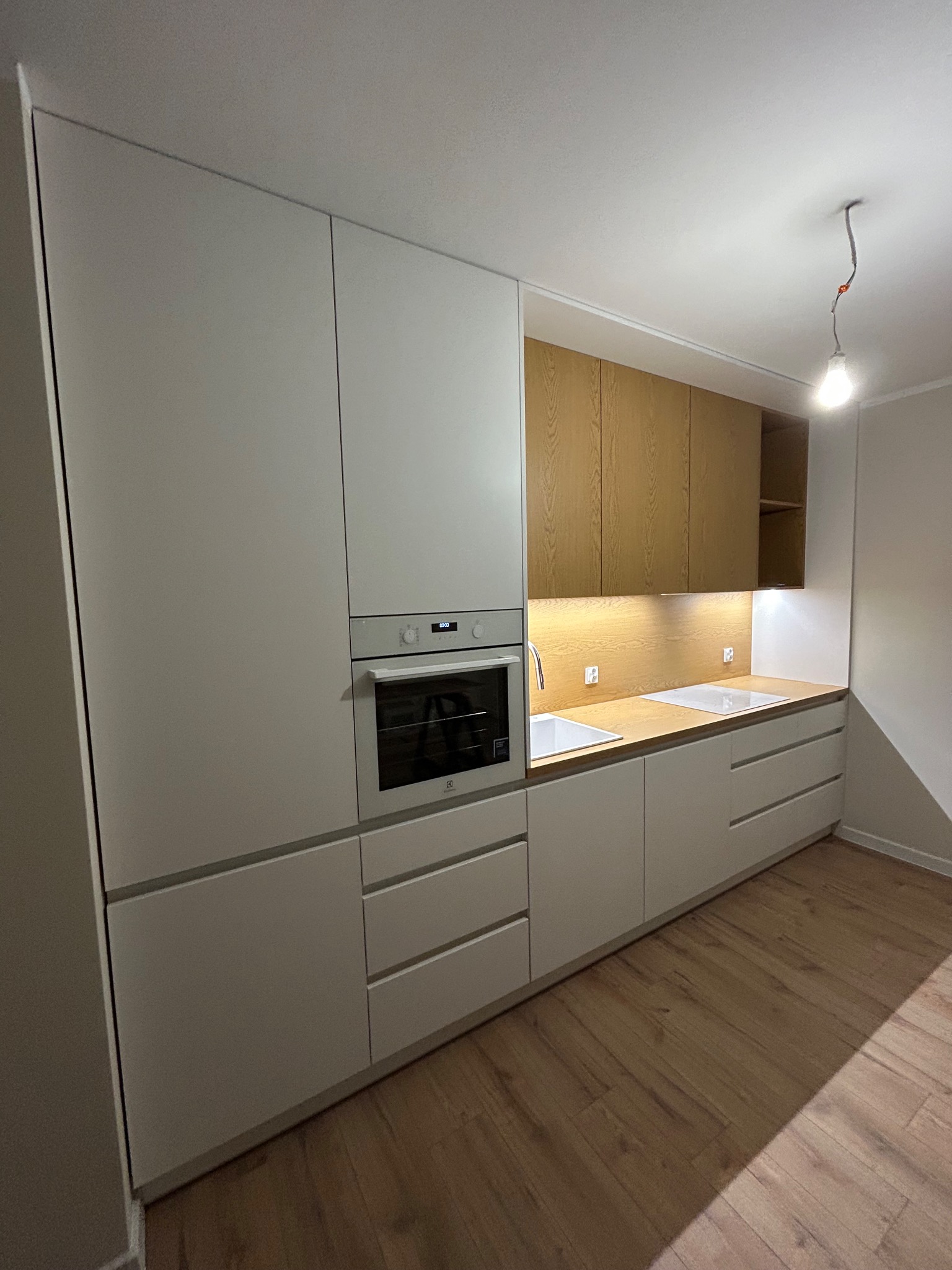 Nowoczesna kuchnia z białymi frontami i drewnianymi szafkami górnymi, oświetlona punktowo. Widoczny piekarnik w zabudowie i drewniany blat z wbudowanym zlewem.