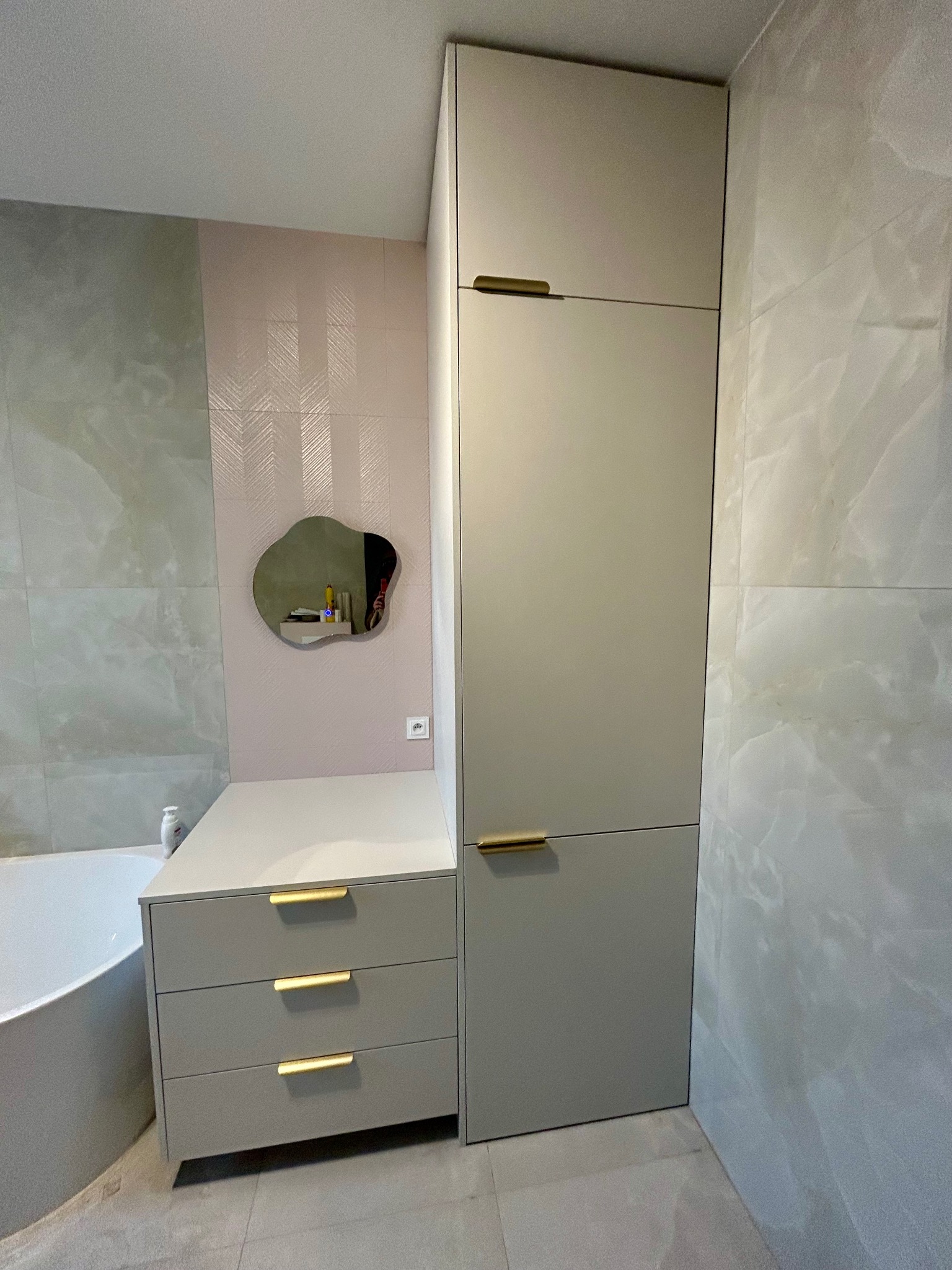 Szara zabudowa łazienkowa z szafkami i lustrem o nieregularnym kształcie na tle marmurowych płytek i różowej ściany z subtelnym wzorem. Złote uchwyty dodają elegancji.