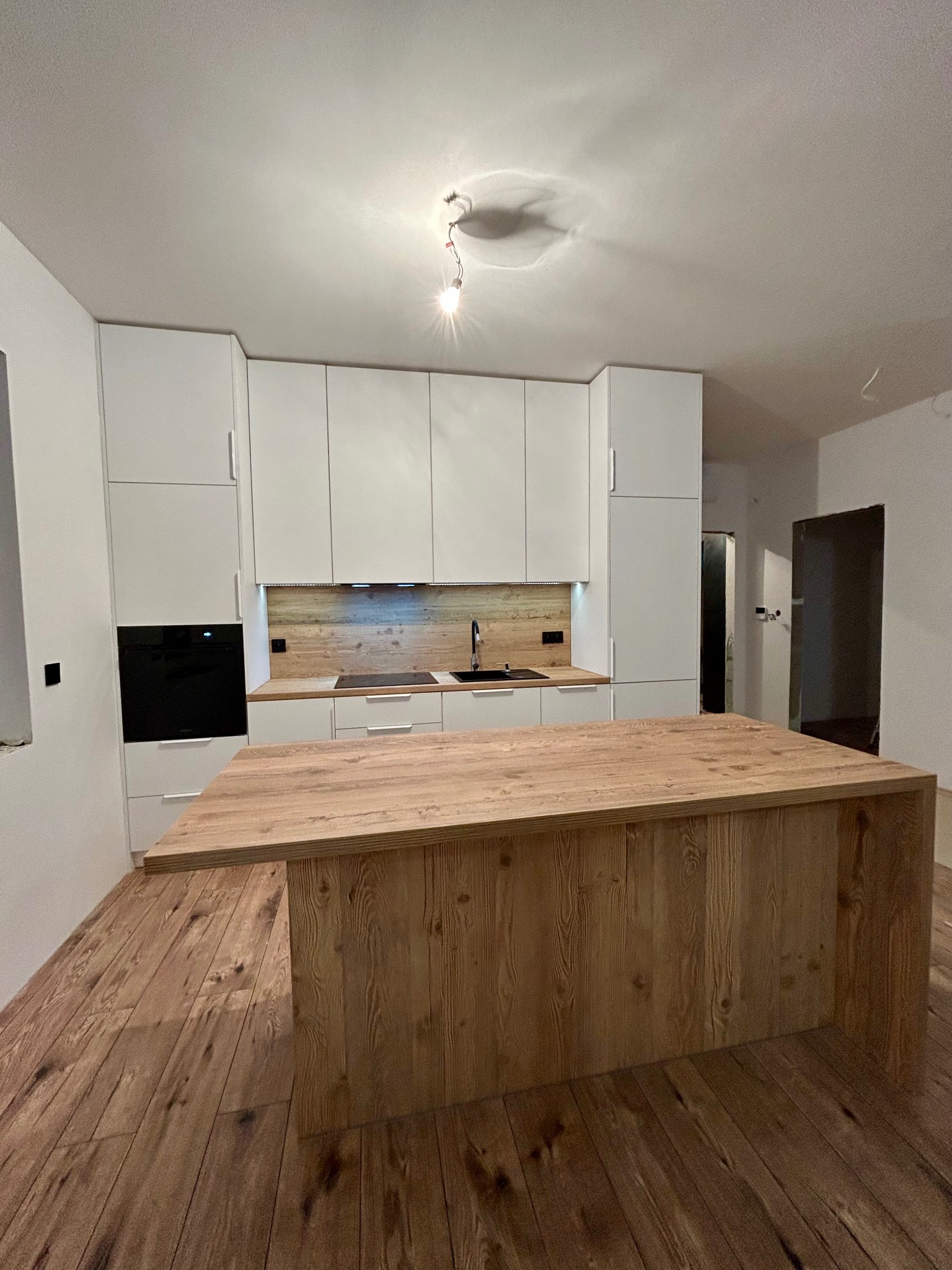 Nowoczesna kuchnia z białymi szafkami i drewnianą wyspą na tle drewnianej podłogi. Widoczne oświetlenie podszafkowe i czarny piekarnik.