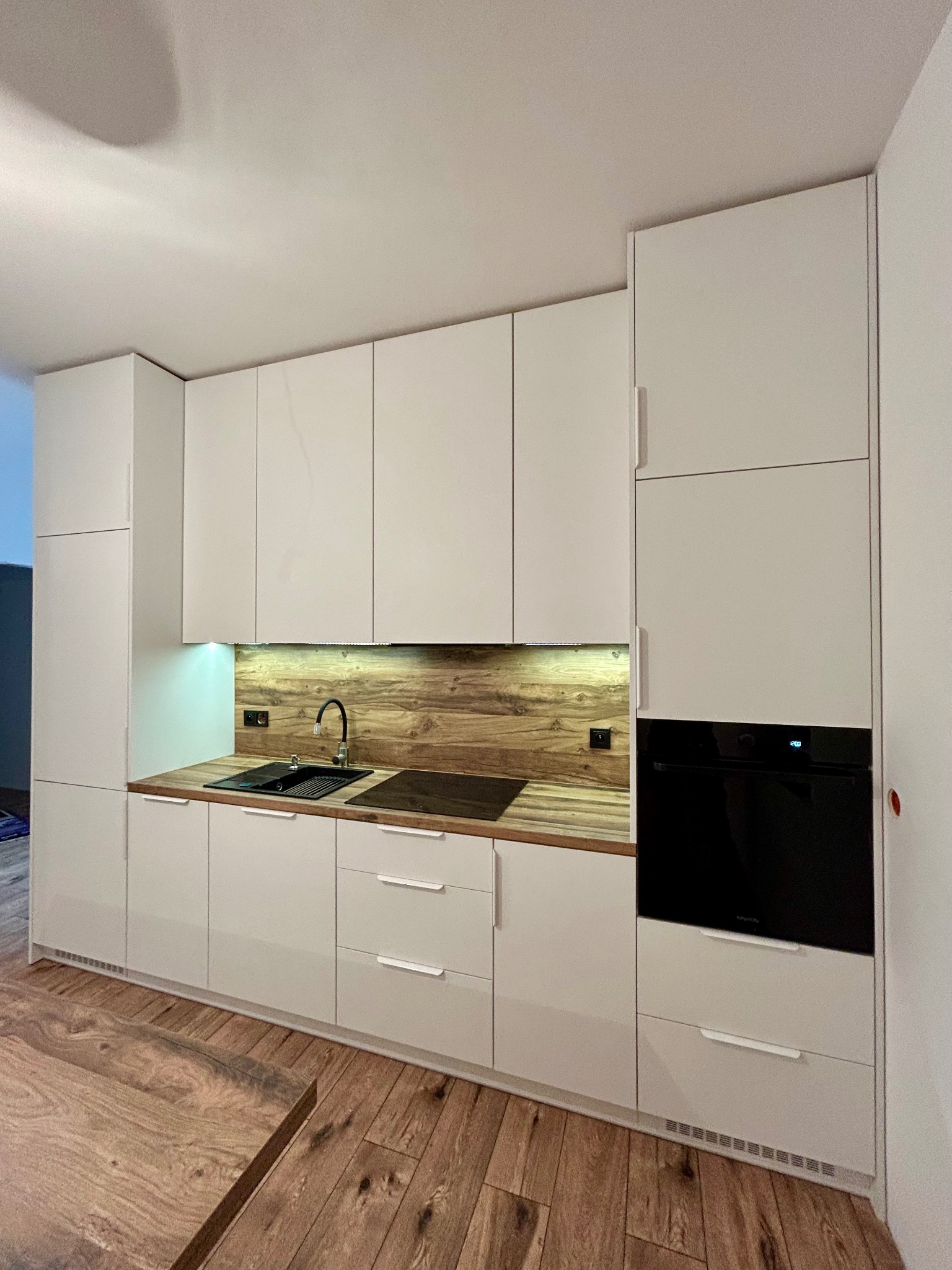 Biała, nowoczesna kuchnia z drewnianym blatem i oświetleniem LED. Uwagę przyciąga czarny piekarnik w zabudowie i drewniana podłoga. Minimalistyczny design.