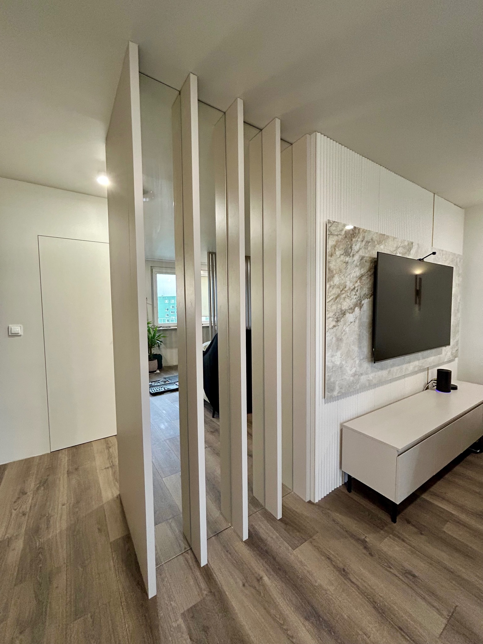 Nowoczesny salon z drewnianą podłogą i ażurową ścianką działową z białych paneli. Telewizor na ścianie z marmurowym akcentem. Minimalistyczny design.