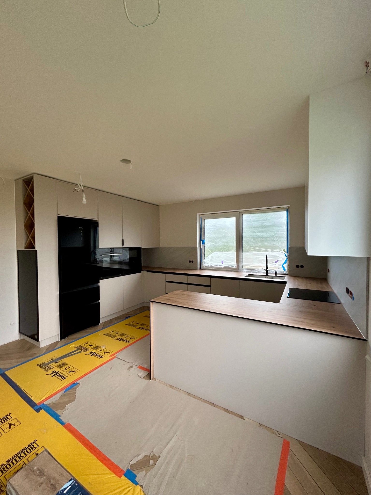 Jasna kuchnia z wyspą w trakcie montażu, zabezpieczona podłoga, widoczne okno i czarny sprzęt AGD. Ujęcie z szerokim kątem.