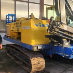 Żółto-niebieska maszyna na gąsienicach z dźwigiem Fassi, widoczna na tle hali z wielkimi oknami, pozostawiająca ślady na betonowym podłożu.