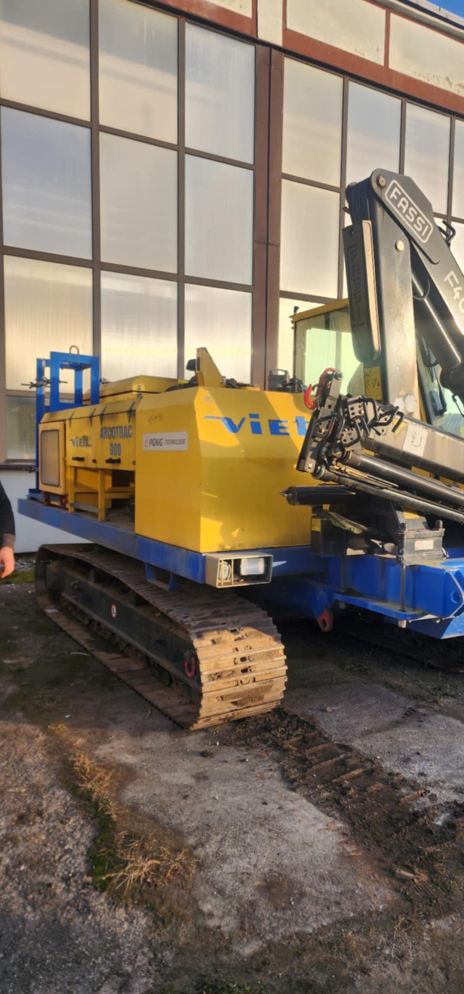 Żółto-niebieska maszyna na gąsienicach z dźwigiem Fassi, widoczna na tle hali z wielkimi oknami, pozostawiająca ślady na betonowym podłożu.