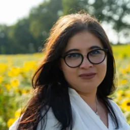 Portret kobiety w okularach na tle pola słoneczników w słoneczny dzień, ubrana w białą koszulę.