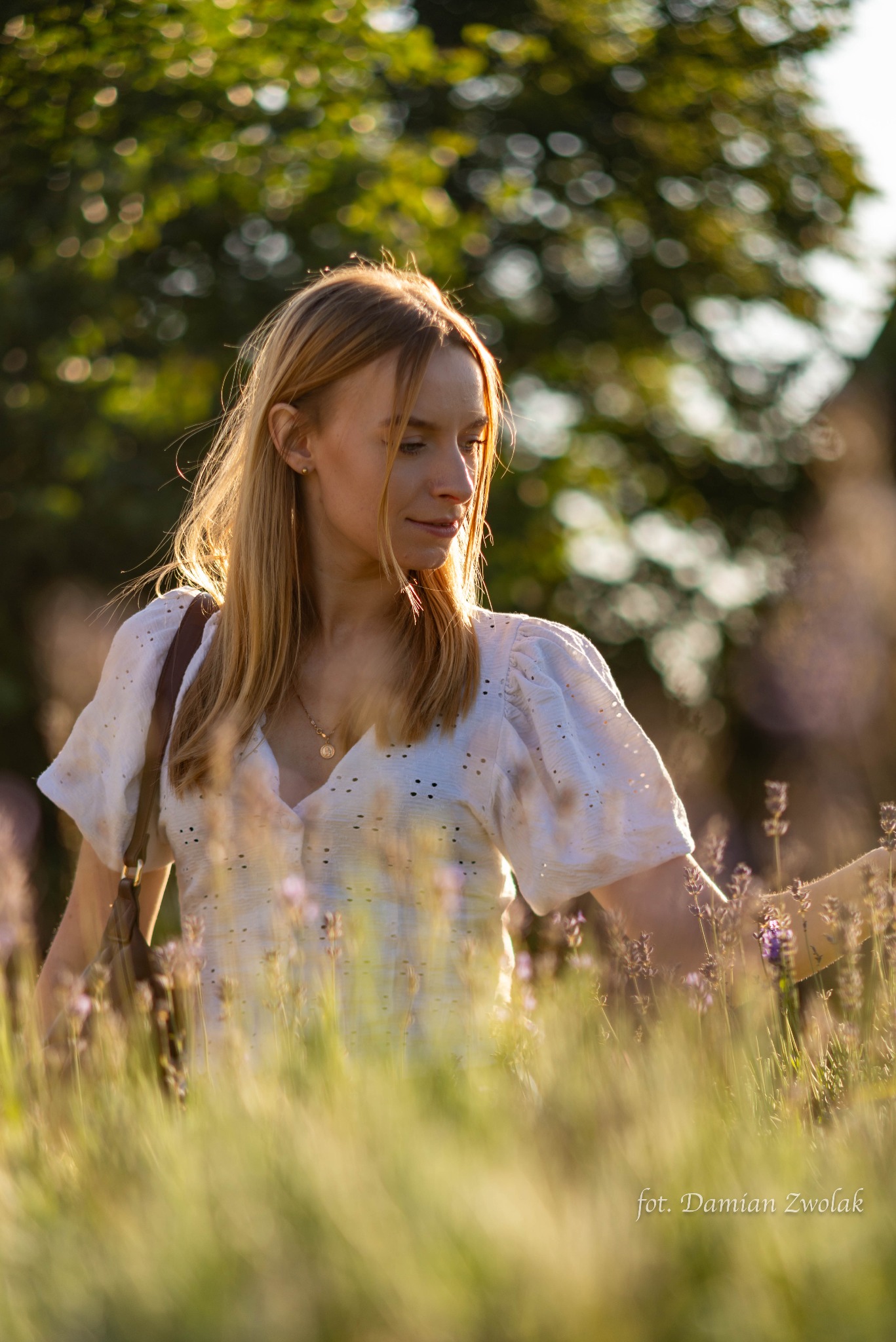 Portret kobiety w białej bluzce z ażurowymi rękawami, stojącej w polu lawendy w promieniach zachodzącego słońca, z brązową torebką na ramieniu i delikatnym złotym naszyjnikiem