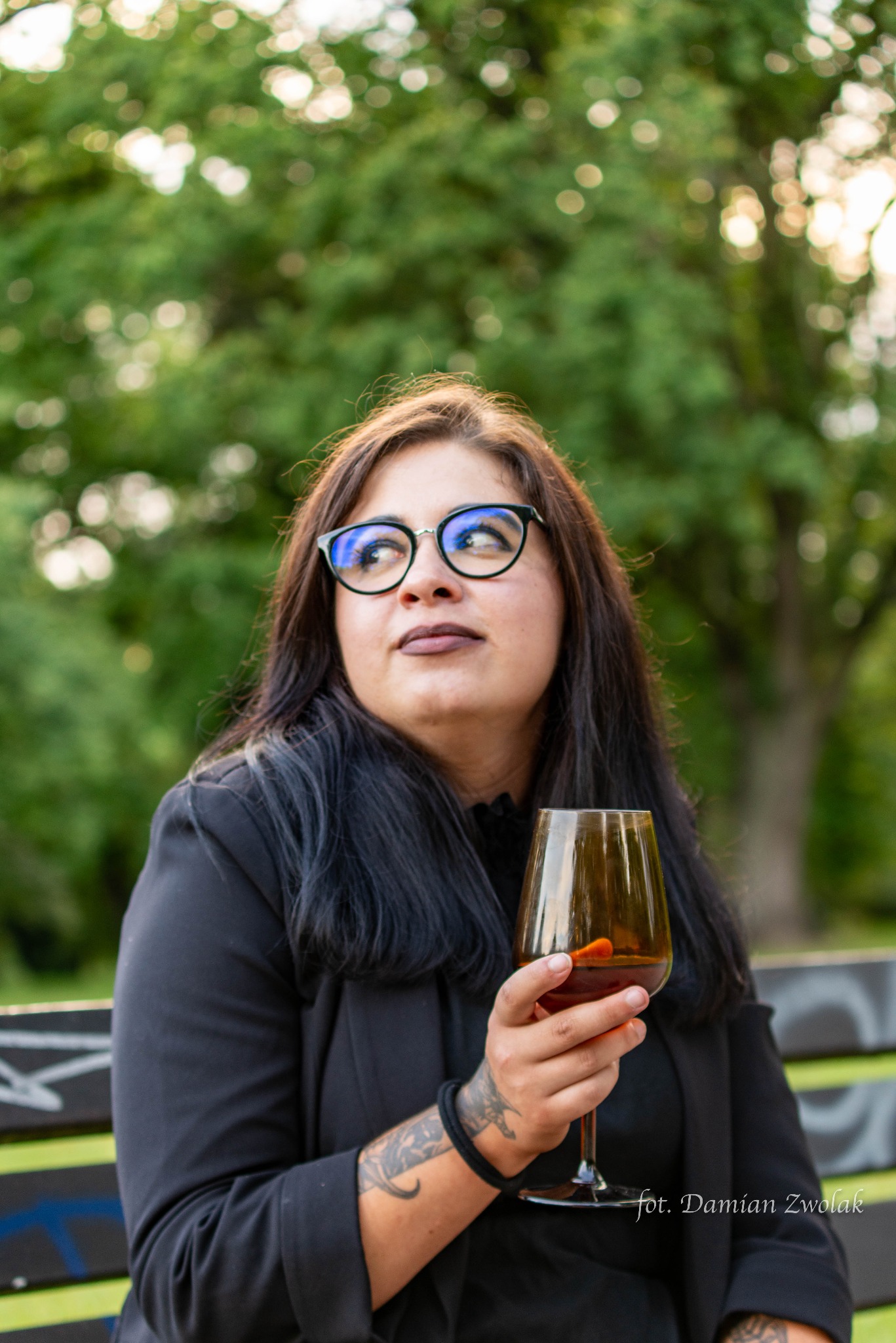 Kobieta w okularach, z tatuażem na ramieniu, trzyma kieliszek z pomarańczowym napojem, siedząc na ławce w parku.