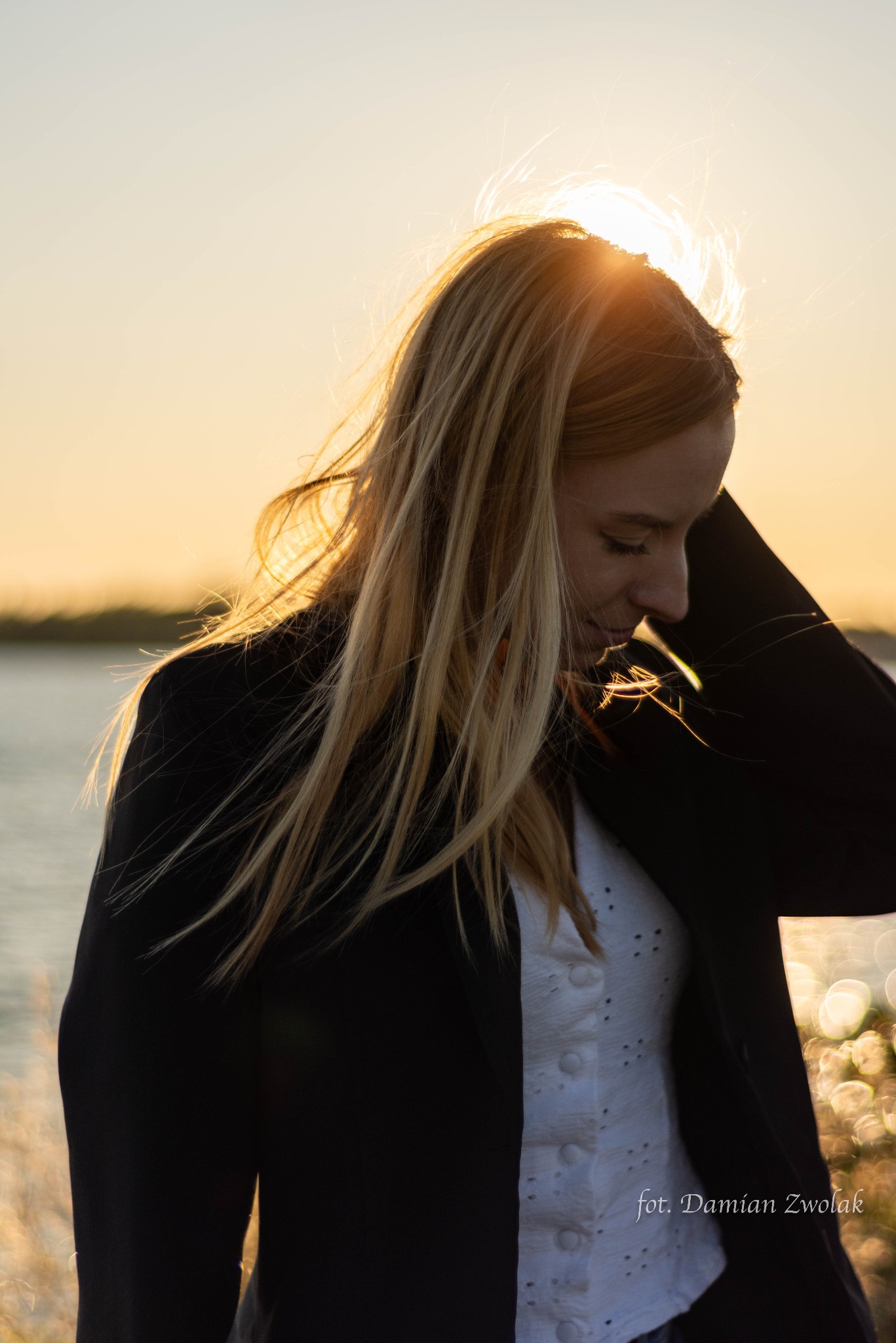 Portret kobiety w czarnej marynarce i białej bluzce na tle zachodzącego słońca nad wodą, z autografem fotografa w prawym dolnym rogu.
