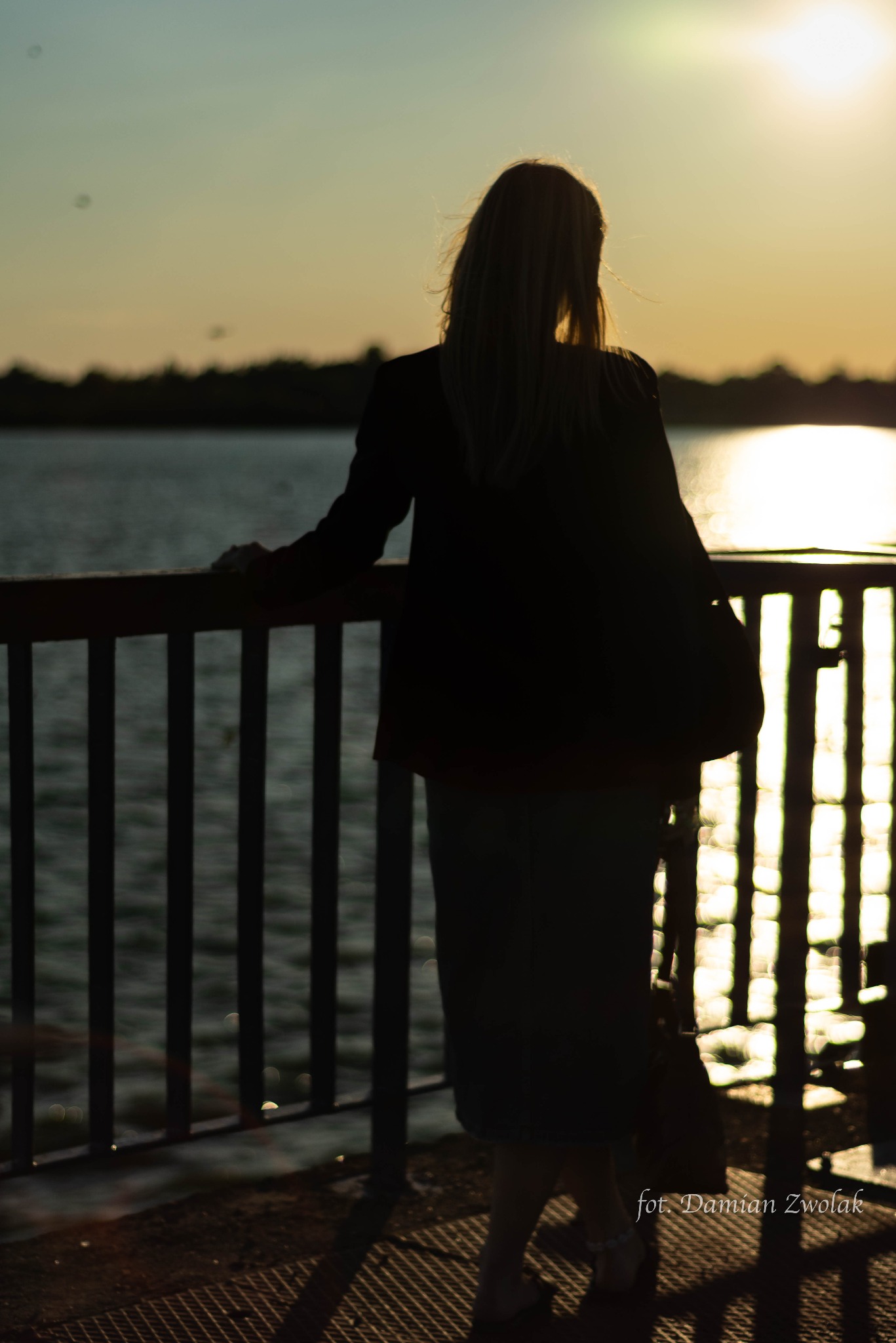 Sylwetka kobiety w marynarce stojącej przy barierce nad wodą o zachodzie słońca, widoczne refleksy na wodzie i delikatne światło na włosach.