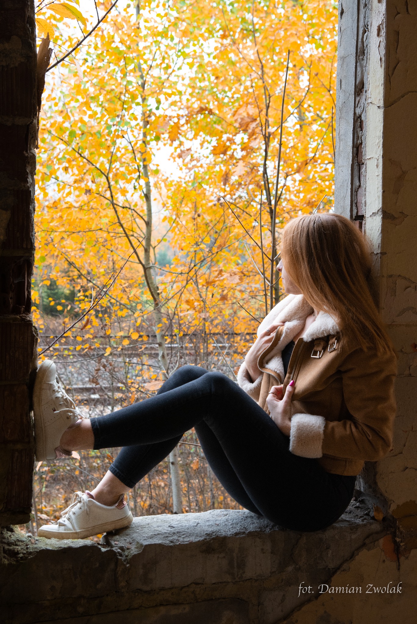 Kobieta siedzi w zniszczonym oknie, ubrana w kurtkę z futerkiem, spoglądając na jesienny las z żółtymi liśćmi.